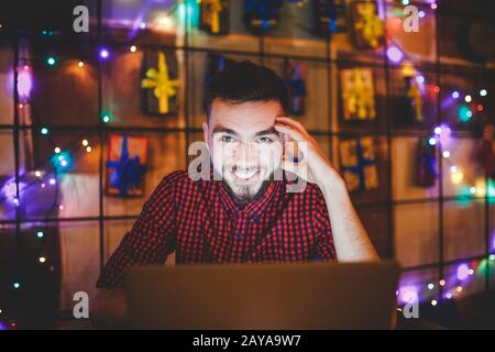 Un giovane bell'uomo caucasico con barba e un sorriso toothy in una camicia rossa a scacchi sta lavorando dietro un notebook grigio seduto a un Foto Stock