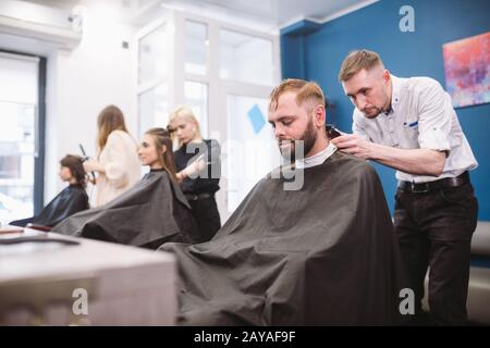 Primo piano di uomo che ottiene haircut trendy al barbiere negozio. Hairstylist maschile che serve il cliente, facendo taglio di capelli con macchina e co Foto Stock