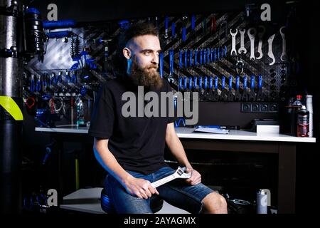 Ritratto di piccola impresa proprietario di giovane uomo con barba. Ragazzo di bicicletta meccanico officina lavoratore seduto con l'attrezzo in mano Foto Stock