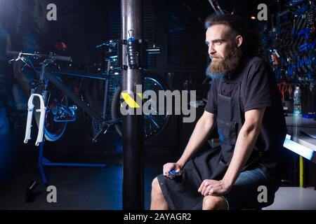 Ritratto di piccola impresa proprietario di giovane uomo con barba. Ragazzo di bicicletta meccanico officina lavoratore seduto con l'attrezzo in mano Foto Stock