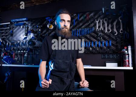 Ritratto di piccola impresa proprietario di giovane uomo con barba. Ragazzo di bicicletta meccanico officina lavoratore seduto con l'attrezzo in mano Foto Stock