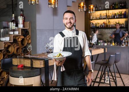 Un bel ragazzo giovane con una barba vestita in un grembiule in un ristorante e con un piatto bianco con una falda. Contro Th Foto Stock