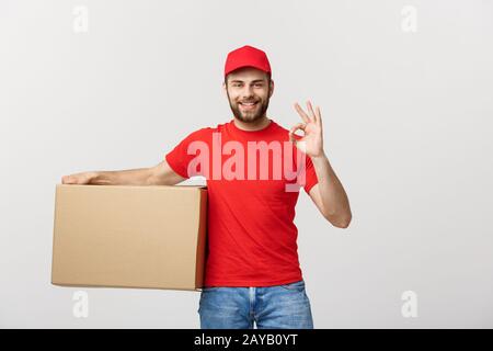 Allegro giovane uomo di consegna nel cappuccio rosso in piedi con pacco postale box isolate su sfondo bianco che mostra bene gesto. Foto Stock