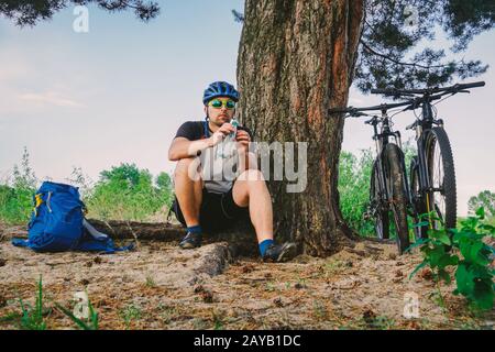 Ciclista maschile caucasica stile di vita attivo riposato sotto l'albero dopo l'esercizio di bicicletta bere acqua dalla bottiglia. Sportivo in hel Foto Stock