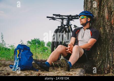 maschio mountain biker appoggiato su un giro in bicicletta, seduto a terra sotto un albero con la sua mountain bike, si trova accanto a lui, godendo di th Foto Stock