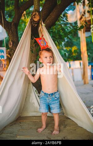 Vacanza estiva a tema, ragazzino, bambino caucasico che gioca in zona boscosa nel parco giochi nel cortile. Il bambino nella parrucca Tipi Foto Stock