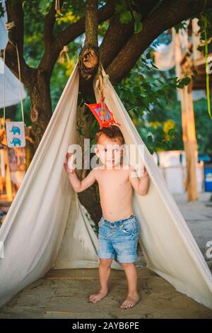 Vacanza estiva a tema, ragazzino, bambino caucasico che gioca in zona boscosa nel parco giochi nel cortile. Il bambino nella parrucca Tipi Foto Stock