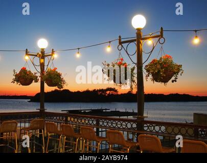 concetto di vacanza scena crepuscolo con sedie da bar intorno luci scintillanti al tramonto su un lago con isola e molo in forma Foto Stock