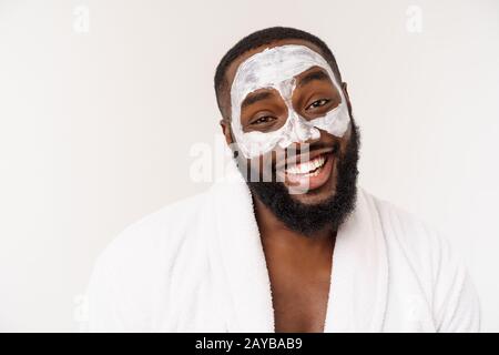 Giovane tipo afro-americano che applica crema viso su sfondo bianco. Ritratto di un giovane felice uomo africano sorridente in studio. Hig Foto Stock