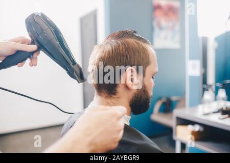 giovane uomo bearded che ottiene cured il parrucchiere con il barbershop dell'asciugacapelli. Parrucchiere Professionale Essicazione Dei Capelli Cliente Bello. Foto Stock