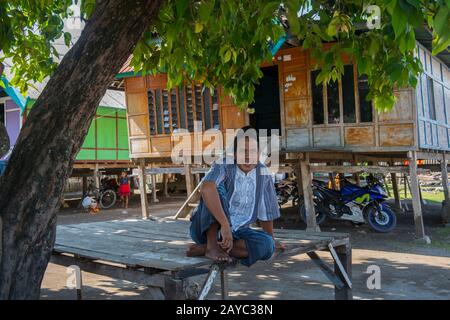 Una scena di villaggio con un uomo seduto di fronte a una casa tradizionale su palafitte nel piccolo villaggio di Moyo Labuon sull'isola di Moyo, al largo della costa di Sumb Foto Stock