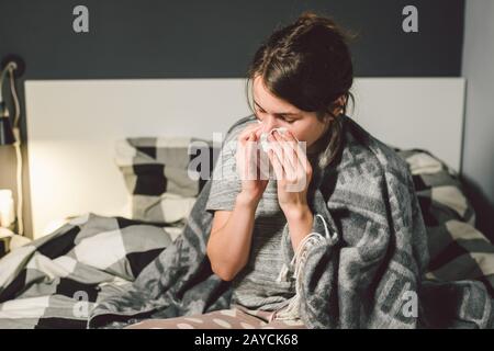donna malata con naso che cola, seduta a letto, malessere e starnutire la camera da letto. Donna che tiene il tessuto, sintomi di influenza. Healthc Foto Stock
