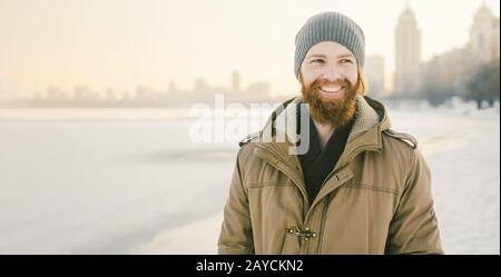 Primo piano giovane caucasico capelli rossi maschio e barba in un cappello e un cappotto parco che posa modello invernale contro uno sfondo di un lago a. Foto Stock