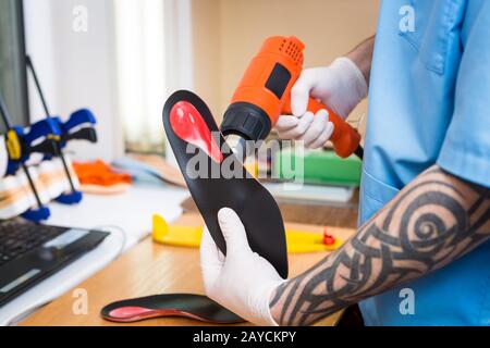 Mani di primo piano di giovane uomo con tatuaggio in laboratorio vestito in uniforme blu fanno solette ortopediche individuali. Lo strumento noi Foto Stock