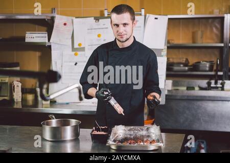 Cucina a tema. Un giovane uomo caucasico in uniforme nera e guanti in lattice in un ristorante in cucina. Minatori di cottura in un essere Foto Stock