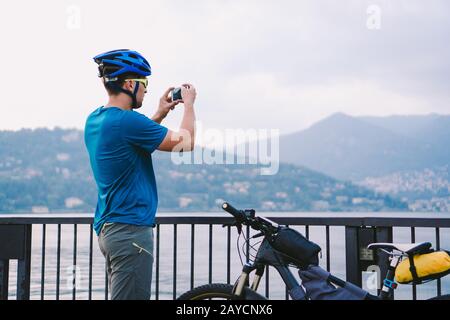 Il ciclista scatta foto con lo smartphone. Uomo caucasico in casco in bicicletta scattare foto del paesaggio. Attività all'aperto. Marca turistica Foto Stock