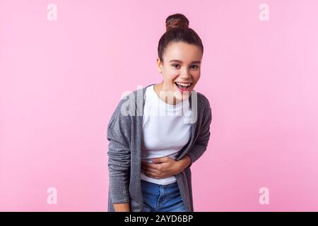 Ritratto di ragazza giovane bruna cute con acconciatura bun in abiti casual scoppiare a ridere, tenendo il suo ventre, guardando gioiosa spensierata divertito Foto Stock
