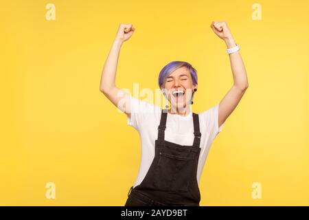 Ritratto di ragazza estatica hippster gioiosa con capelli viola corti in tute denim in piedi con i suoi pugni alzati e gridando con entusiasmo, vivace Foto Stock