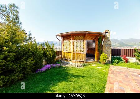 Un gazebo in legno con una stufa a barbecue in pietra nel cortile del cottage. Gazebo accanto alla recinzione nell'angolo del cortile. C'è un prato verde Foto Stock