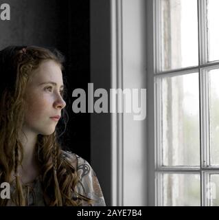 Una ragazza dai capelli rossi con frane e trecce, in luce soffusa, guardando attraverso una finestra fuori in lontananza. Foto Stock