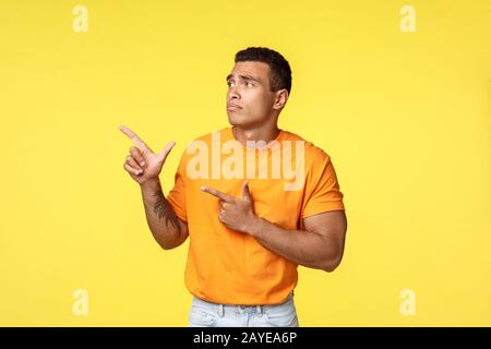 Geloso e sconvolto uomo carino maschile, indossare t-shirt arancione, in piedi infelice e triste su sfondo giallo, punto di spoloramento Foto Stock
