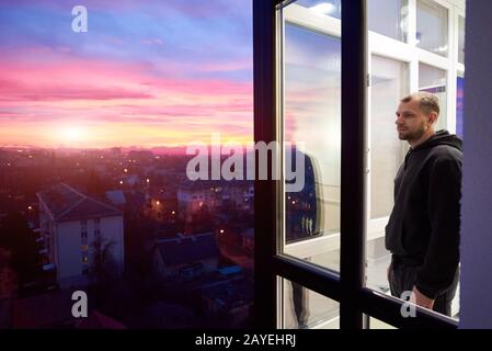 Vista laterale di un uomo in piedi su un balcone con finestre panoramiche e guardando in lontananza mentre godendo il magico tramonto. Paesaggio urbano dall'altezza e un cielo di fuoco multicolore con tramonto Foto Stock