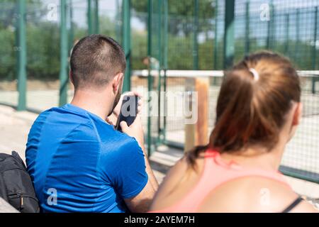 Uomo con una barba vestita di abbigliamento sportivo seduto su un campo da tennis utilizzando il cellulare con una ragazza accanto a lui Foto Stock