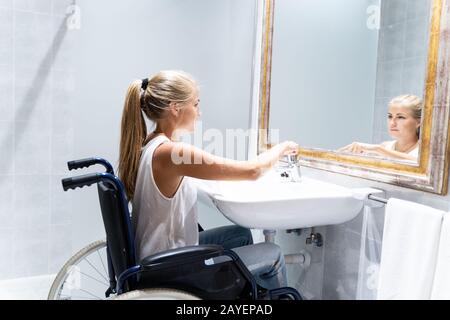 Ragazza bionda in sedia a rotelle con capelli lunghi che girano sul rubinetto d'acqua in un bagno di fronte allo specchio Foto Stock