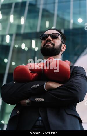 Foto verticale di un uomo d'affari sorridente con occhiali da sole e guanti da boxe rossi appesi al collo con le braccia incrociate davanti a un edificio Foto Stock