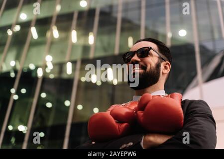 Uomo d'affari sorridente con occhiali da sole e guanti da boxe rossi appesi al collo con le braccia incrociate davanti a un edificio Foto Stock