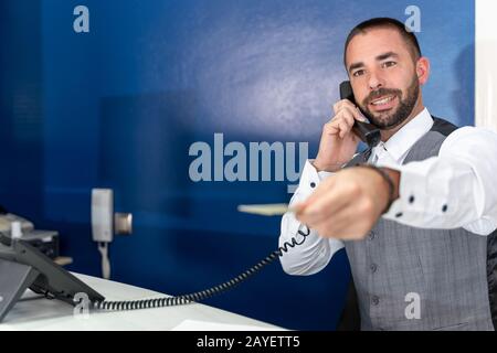 Foto di scorta di un receptionist dell'hotel che parla al telefono con un'espressione amichevole mentre estende il suo braccio con una scheda della stanza nella sua mano Foto Stock