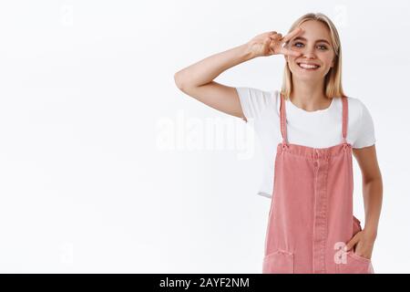 Allegra, giovane e positiva donna bionda moderna in tute rosa, t-shirt, tenere la mano in tasca, mostrare la vittoria o il segno di pace sopra l'occhio, sorridendo toohy Foto Stock