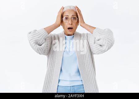 Vecchia signora nervosa e travagliata con capelli pettinati grigi, occhiali di protezione e abiti comodi, afferrare la testa in mani da ansia e paura, gasping Foto Stock