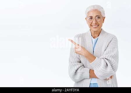Allegro piacevole donna anziana, nonna con capelli pettinati grigi, occhiali, punta in alto a sinistra per promuovere la pubblicità, dare raccomandazione Foto Stock