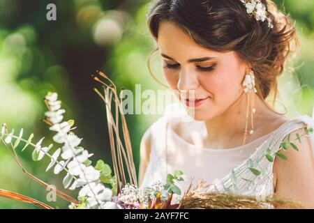 Bella sposa con fiori di bouquet si erge sullo sfondo della foresta. Stile rustico. Bellissima sposa in abiti delicati all'aperto. Clos Foto Stock