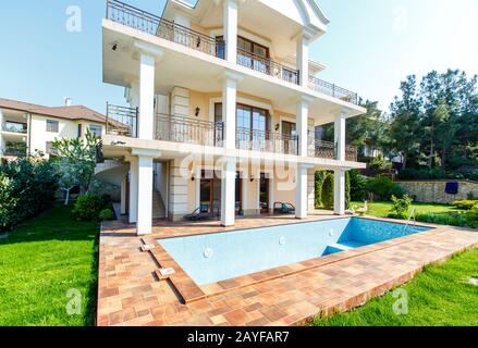 Il cortile posteriore di un cottage bianco a tre piani con colonne e ringhiere in ferro battuto, stile italiano, classico. Giardino verde, prato e piscina vuota in fron Foto Stock