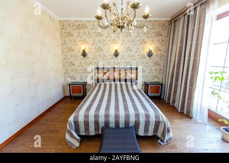 Elegante camera da letto costosa con un grande letto bianco con un copriletto a righe. Un'alta testata con un motivo rosso, lampade a parete intrecciate e un lampadario. B Foto Stock