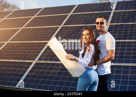 Giovane coppia dolce sorridente è in piedi da un modulo solare in una giornata di sole, guardando la fotocamera, tenendo un piano di carta indossando abiti simili, copiare spazio, vista laterale Foto Stock