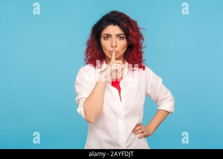 Shi, fai silenzio! Ritratto di donna hippster dispiaciuto con fantasia capelli rossi in camicia bianca tenendo un dito sulle labbra, arrossendo e mostrando il gesto di silenzio. In Foto Stock