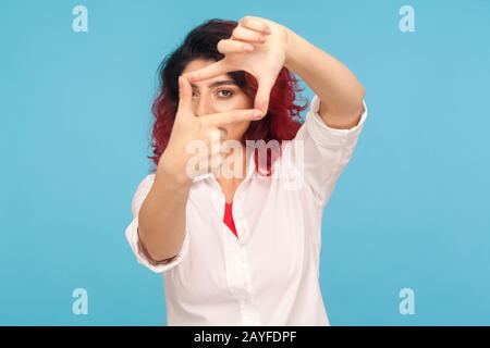 Ritratto di donna con fantasia capelli rossi guardando attraverso la cornice delle dita gesturante foto, zoom messa a fuoco alla fotocamera, osservando con seria espressione. Foto Stock