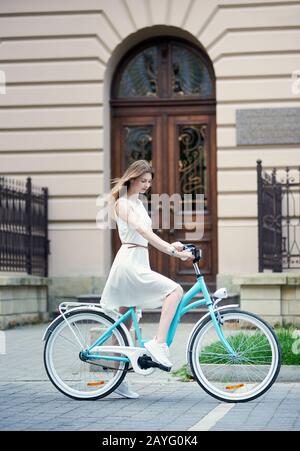 Bella ragazza in un abito leggero e sneakers su una bici blu retrò sullo sfondo di un vecchio edificio con una bella porta. Gite in bicicletta nel pomeriggio estivo all'aperto Foto Stock