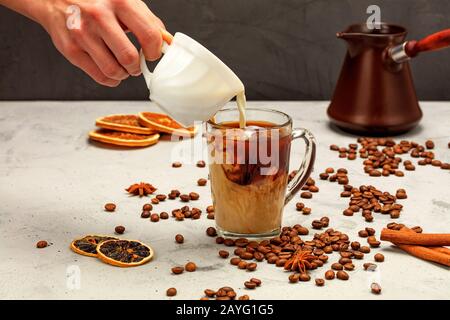 In un espresso appena fatto in una tazza di vetro trasparente, una mano dell'uomo aggiunge la crema bianca, circondata da chicchi di caffè, fette essiccate di arancia e limone. Foto Stock