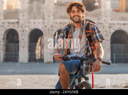 Felice giovane turista in Bicicletta Equitazione indossando maglietta e hat al Colosseo a Roma, Italia di sunrise. Foto Stock