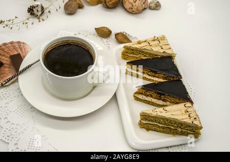 Una tazza bianca con caffè nero e vari pezzi di torte color crema su un piatto bianco tra noci e conchiglie si trovano su un tovagliolo di carta bianca Foto Stock