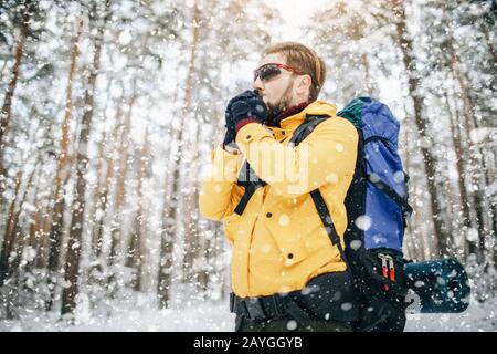 Vista ad angolo basso di giovane uomo barbuto escursionista cercando di ottenere le sue mani calde nella foresta d'inverno Foto Stock