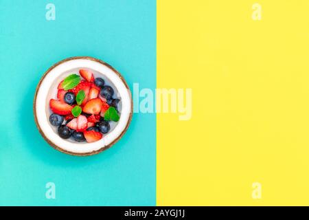 Composizione creativa con ciotola di cocco con frutti di bosco freschi crudi su sfondo geometrico blu e giallo. Vista dall'alto, spazio di copia Foto Stock