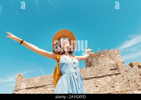 Felice donna turistica in vacanza posa con cappello e zaino di fronte alla vecchia fortezza in rovina e faro a Rodi, Grecia Foto Stock