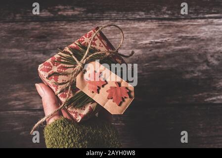 Tenendo una mano un piccolo regalo segreto di Natale con creativo dono rustico decorativo fai da te avvolto in carta retrò rosso da imballaggio Foto Stock