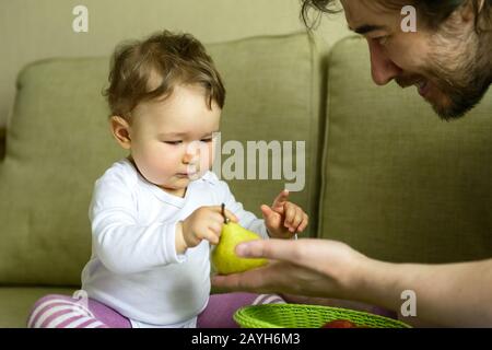 La ragazza cute del bambino gioca con i frutti con il suo padre a casa. Il bambino di un anno guarda attentamente la pera. Foto Stock