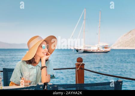 Donna felice che indossa un grande cappello seduto in un bar e che guarda al mare Foto Stock